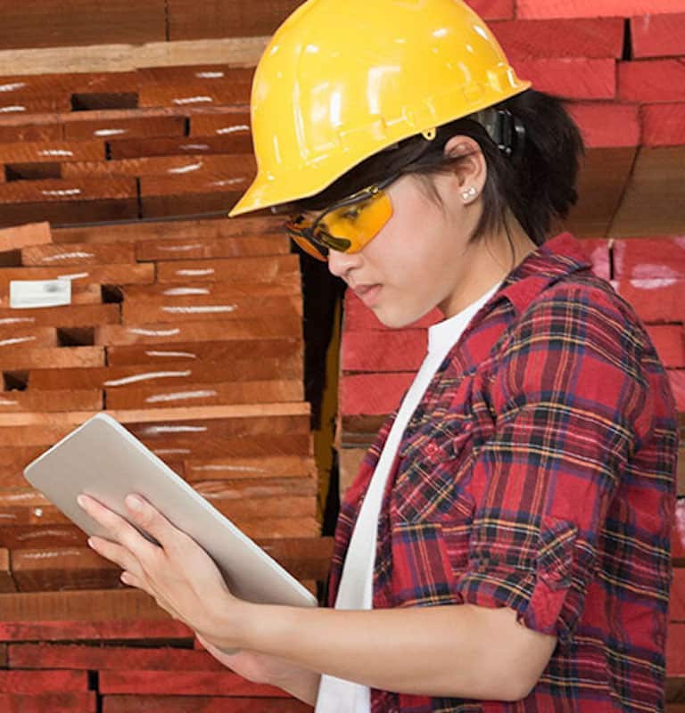 woman in a plaid shirt and a yellow hard hat checking a tablet in a warehouse filled with shelves of lumber